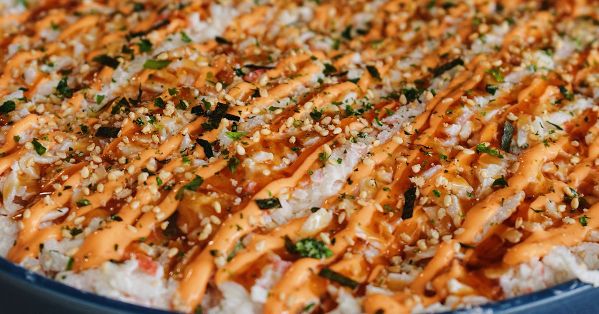 A top-down view of a completed California Roll Sushi Bake served in a large, round navy-blue bowl, heavily covered in spicy mayo, eel sauce, and furikake.