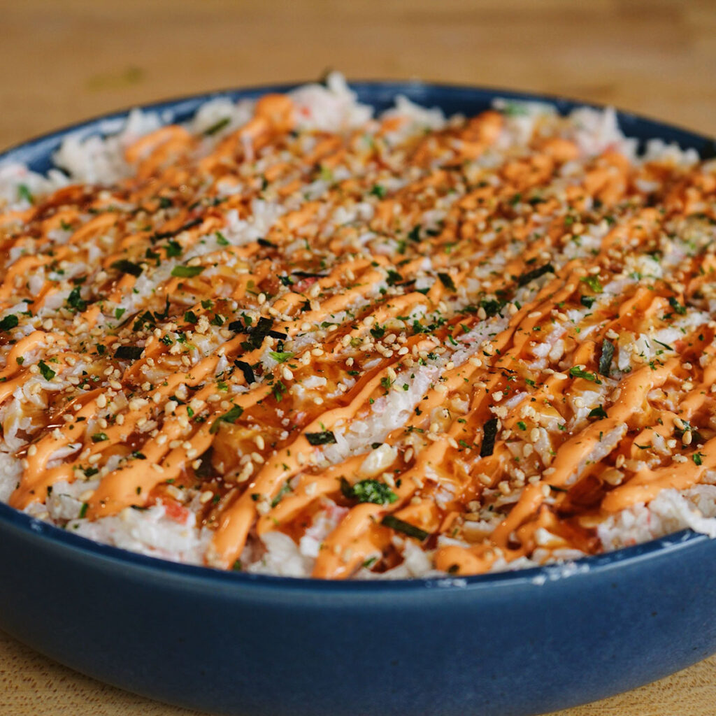 A close-up of a large blue bowl filled with a California Roll Sushi Bake, topped with spicy mayo and eel sauce.