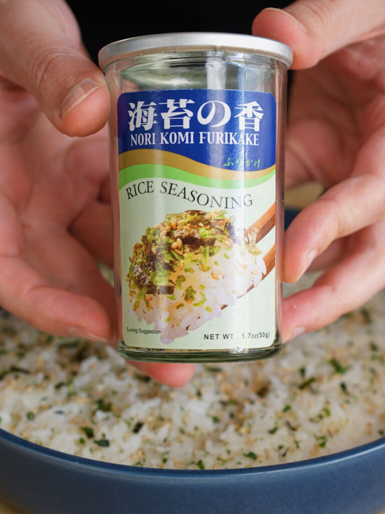 A hand sprinkling Nori Komi Furikake (rice seasoning) over the pressed layer of seasoned sushi rice in a navy-blue bowl for a California Roll Sushi Bake.