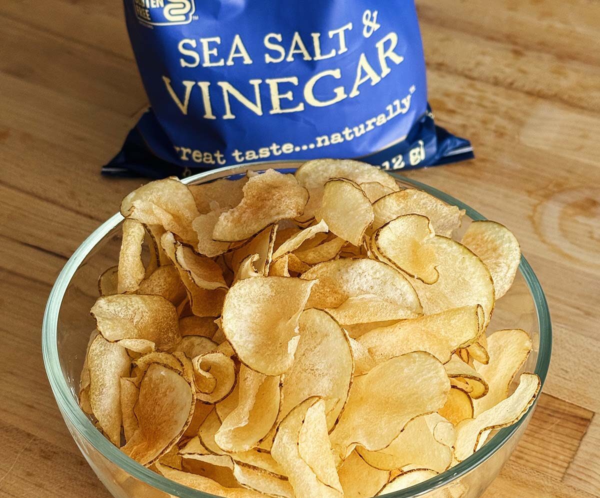 A glass bowl filled with crispy homemade Salt and Vinegar Potato Chips sitting on a wooden cutting board in front of a blue bag of Kettle Brand chips.