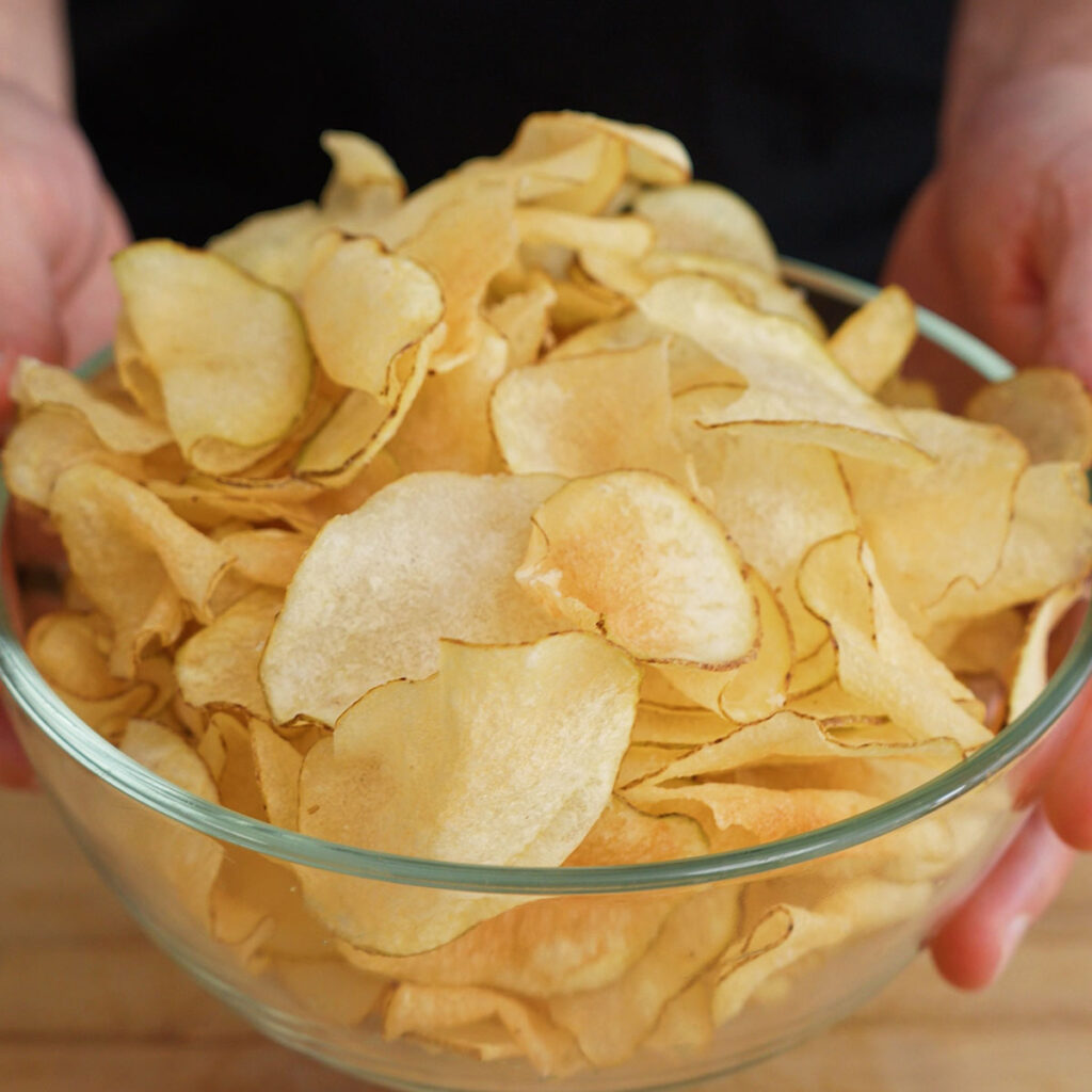 Two hands holding out a large clear glass bowl overflowing with freshly cooked, pale golden homemade Salt and Vinegar Potato Chips.