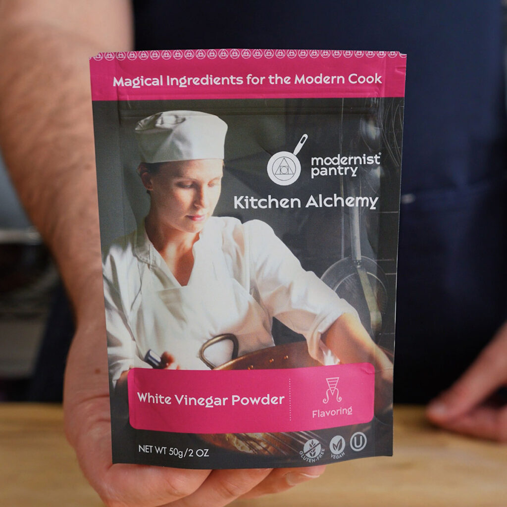 A hand holding up a pouch of Modernist Pantry White Vinegar Powder, the secret dry flavoring ingredient used for homemade Salt and Vinegar Potato Chips.