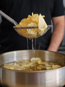 A wire spider skimmer lifting a fresh batch of golden, fully dehydrated Salt and Vinegar Potato Chips out of a pot of hot frying oil.