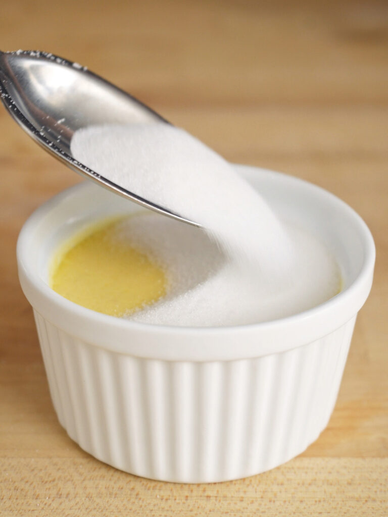 Using a spoon to sprinkle an even layer of white granulated sugar over a chilled homemade creme brulee.