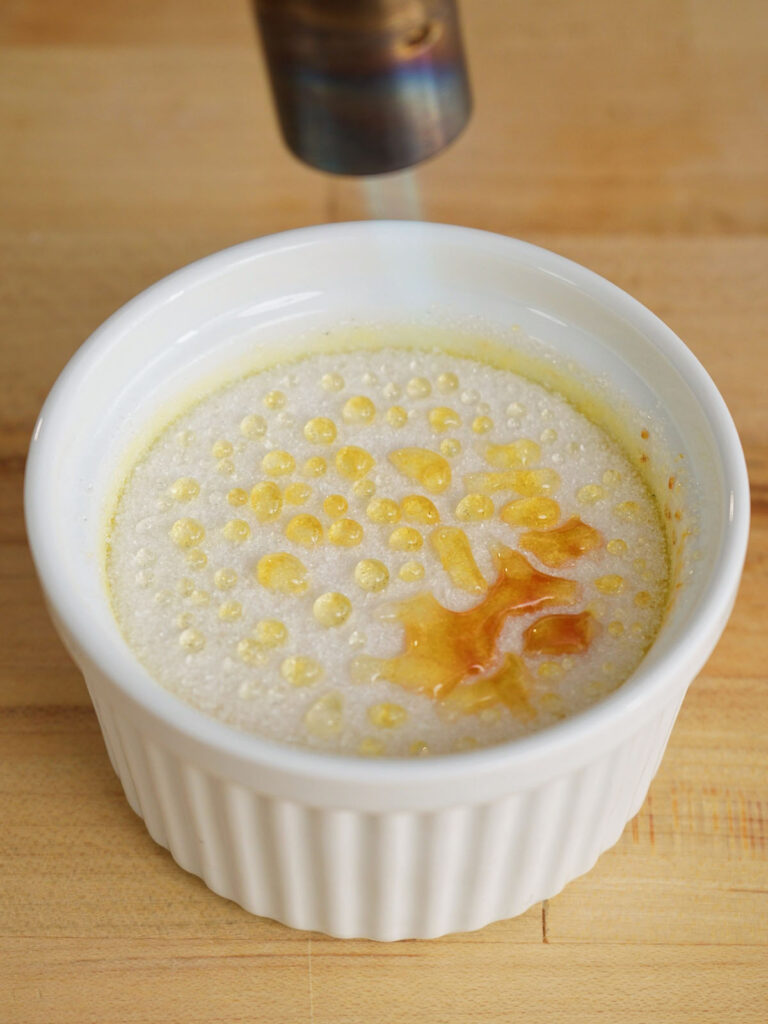 A culinary blowtorch melting and caramelizing the granulated sugar on top of a homemade creme brulee to form a glass shell.