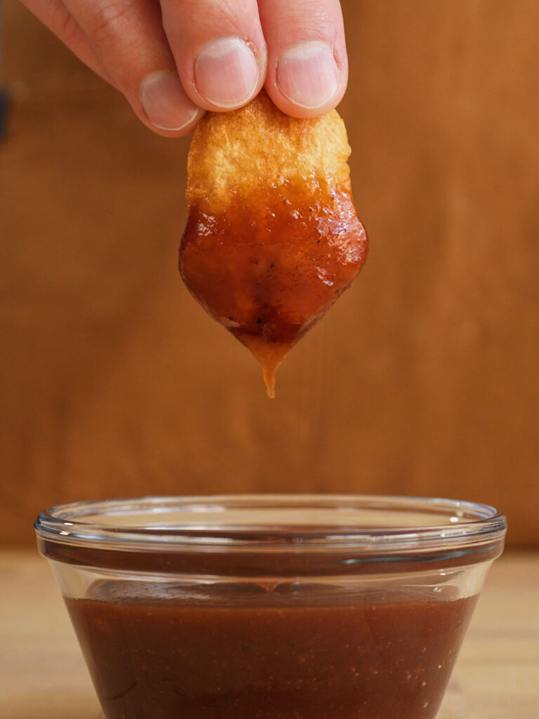 A hand dipping a freshly fried, crispy homemade chicken nugget into a clear glass bowl filled with rich barbecue sauce.