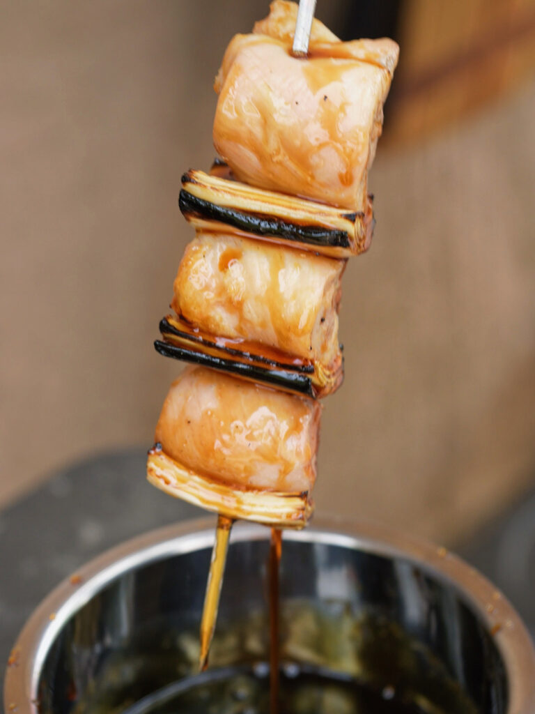 Dipping a partially cooked chicken and charred leek skewer directly into a pot of rich, dark tare sauce for a homemade yakitori recipe.