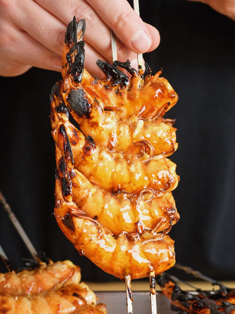 Holding up a double-skewered row of grilled shrimp dripping with sweet and savory tare sauce, freshly cooked for a homemade yakitori recipe.