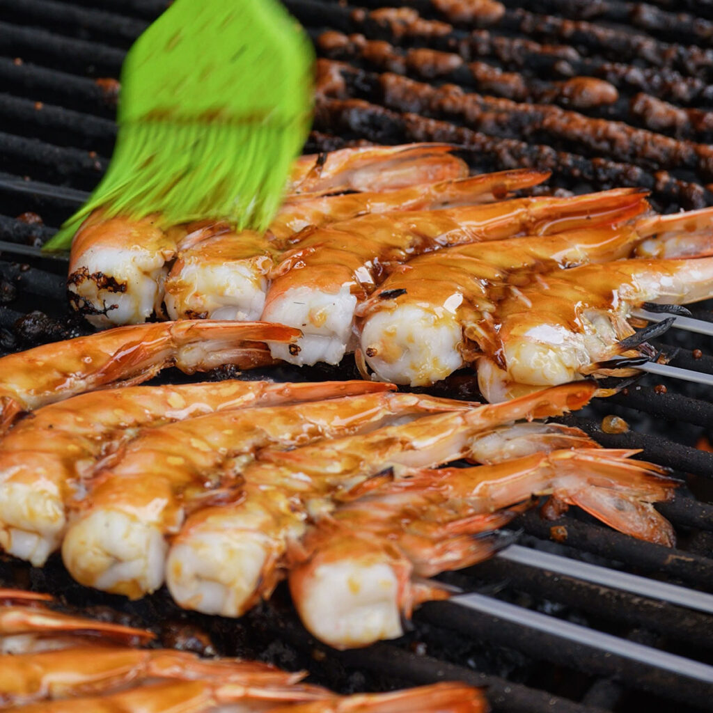 Using a green silicone brush to generously baste fresh shrimp skewers with tare sauce on a hot grill for a homemade yakitori recipe.