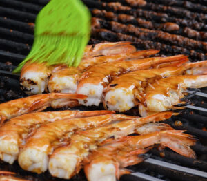 Using a green silicone brush to generously baste fresh shrimp skewers with tare sauce on a hot grill for a homemade yakitori recipe.