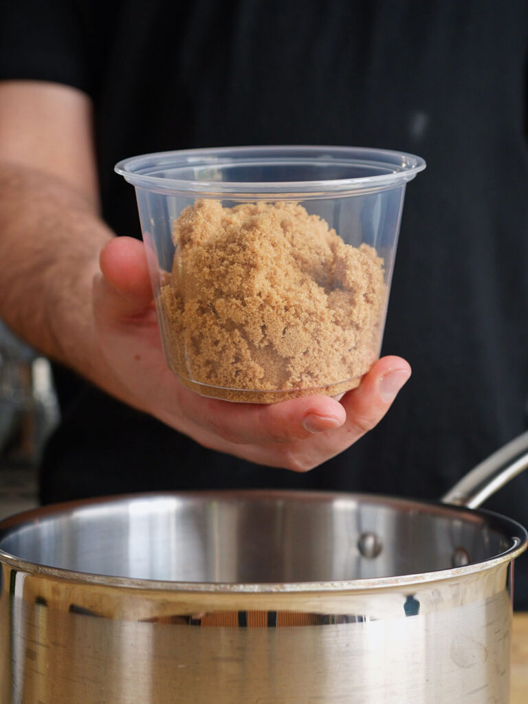 A hand holding a clear container of packed brown sugar over a stainless steel saucepan to make tare sauce for a homemade yakitori recipe.