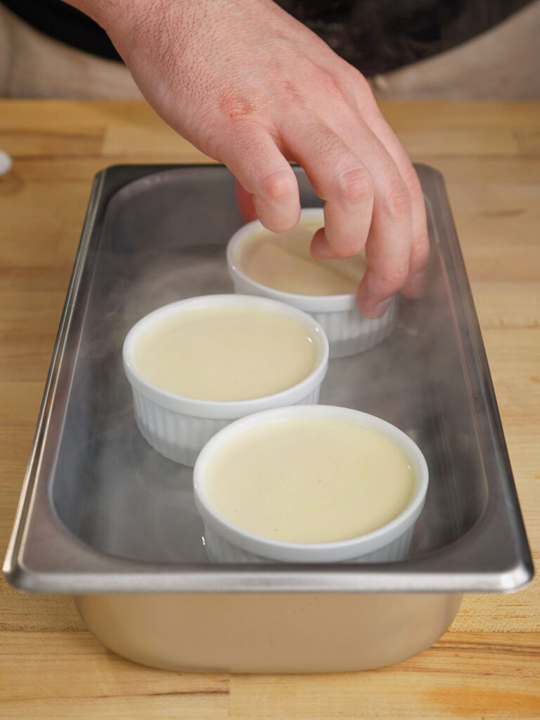 Carefully placing white ceramic ramekins filled with homemade creme brulee custard into a hot water bath.
