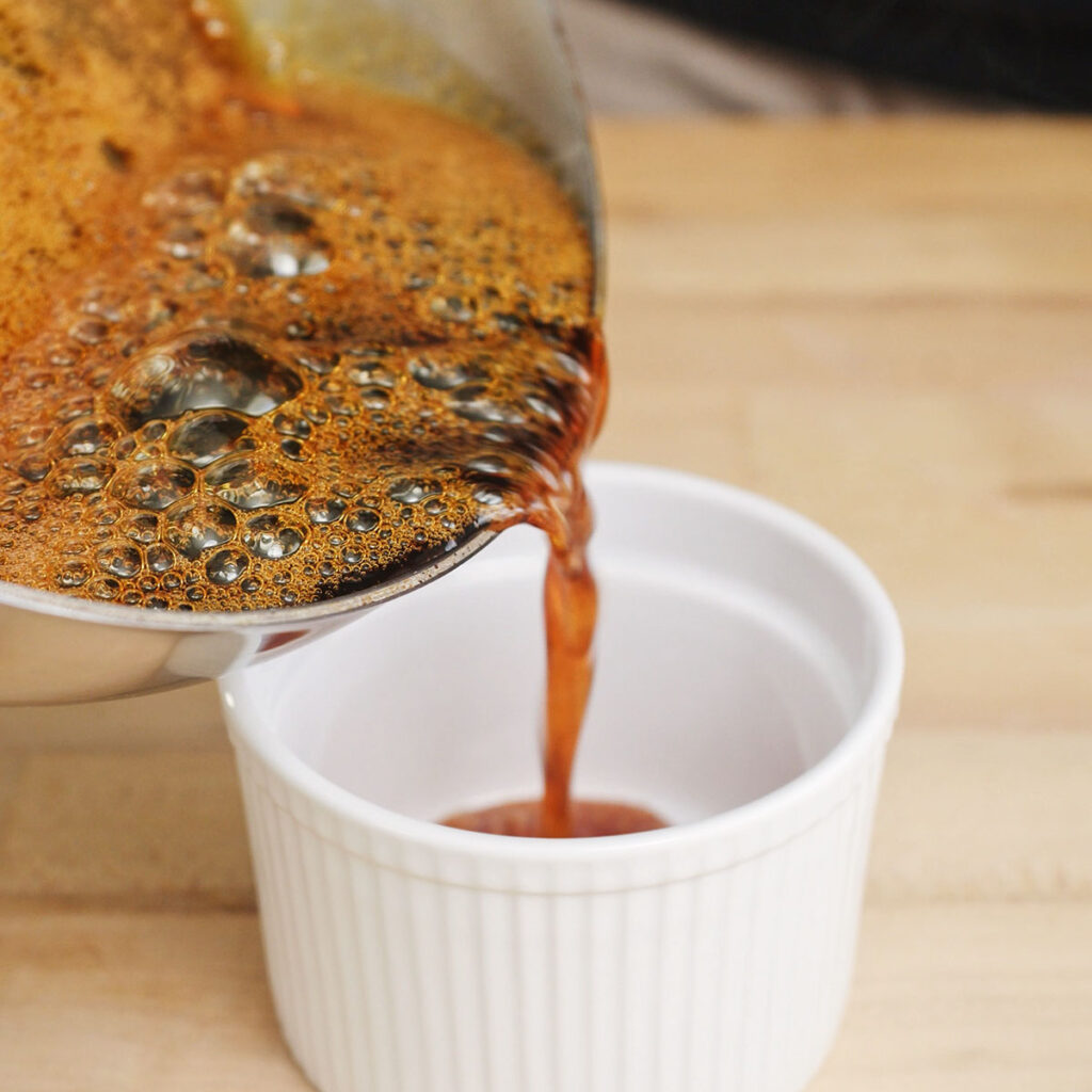 Pouring hot liquid caramel into the bottom of a ceramic ramekin to start making homemade flan.