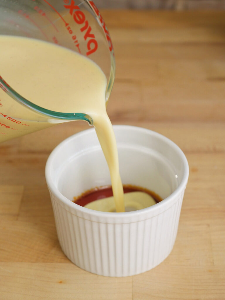 Pouring the strained custard mixture over the hardened caramel inside a ramekin to prepare homemade flan.