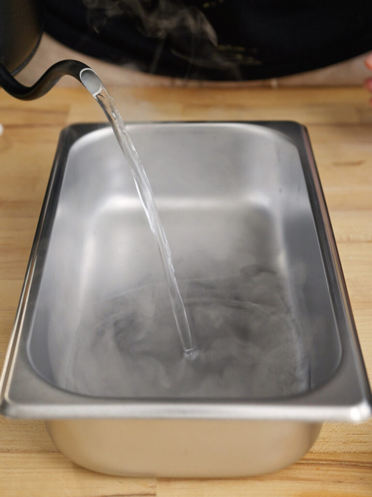 Pouring steaming hot water into a metal roasting pan to create an insulated water bath for baking homemade creme brulee.