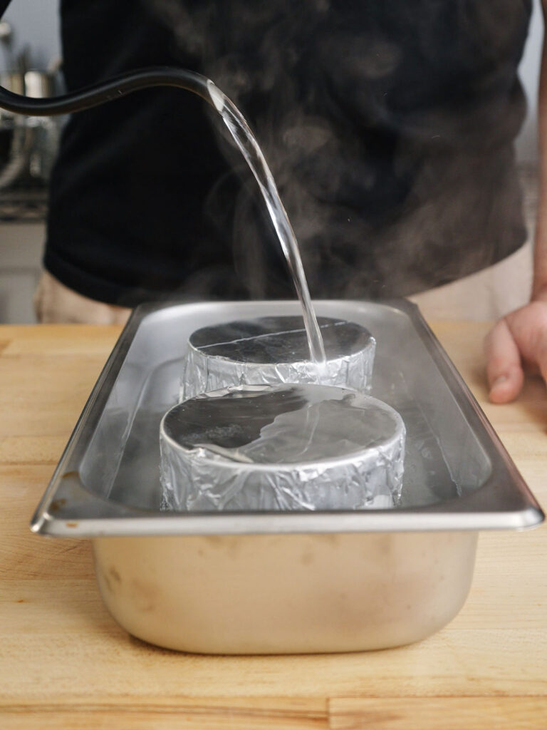 Pouring boiling water into a roasting pan around foil-covered ramekins to create a gentle water bath for baking homemade flan.