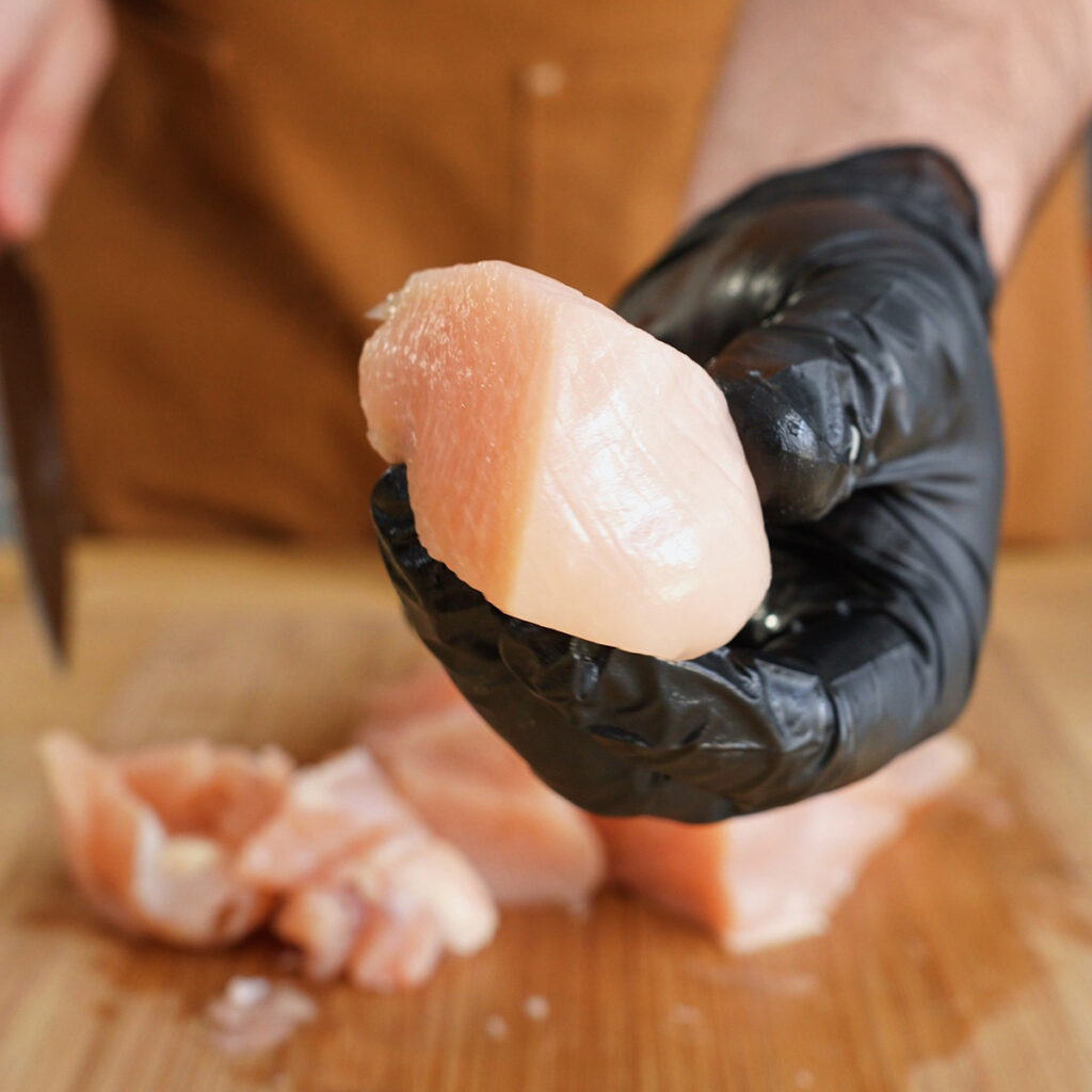 Black-gloved hand holding a thick, fresh piece of raw chicken breast used for making a chicken nugget meat emulsion.
