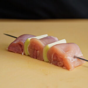 Bite-sized pieces of raw chicken thigh alternating with fresh leeks on a metal skewer, prepped for a homemade yakitori recipe.