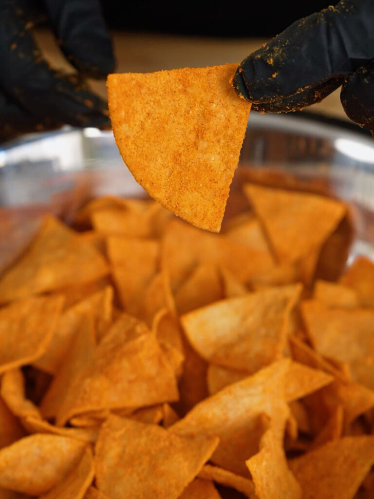 A black-gloved hand holding up a single, perfectly coated DIY Nacho Cheese chip, showcasing the results of Roice Bethel's (@noflakeysalt) food science techniques.