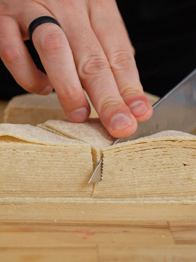 Chef Roice Bethel (@noflakeysalt) slicing a stack of fresh white corn tortillas into triangles to prep for his from-scratch savory chip recipe.
