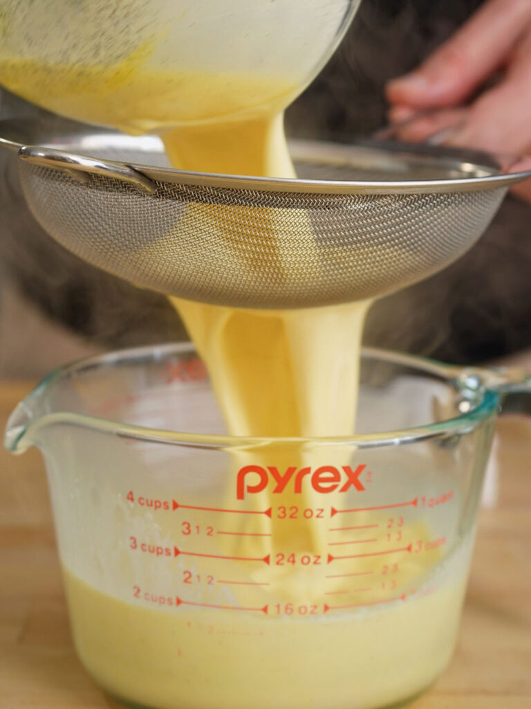 Pouring the liquid homemade creme brulee base through a fine mesh strainer into a glass measuring cup to ensure a silky texture.