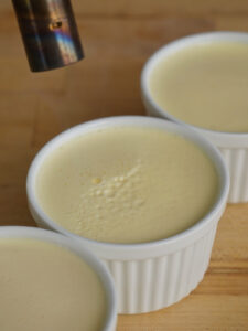 Using a culinary blowtorch to quickly pop surface air bubbles on unbaked homemade creme brulee in white ramekins.
