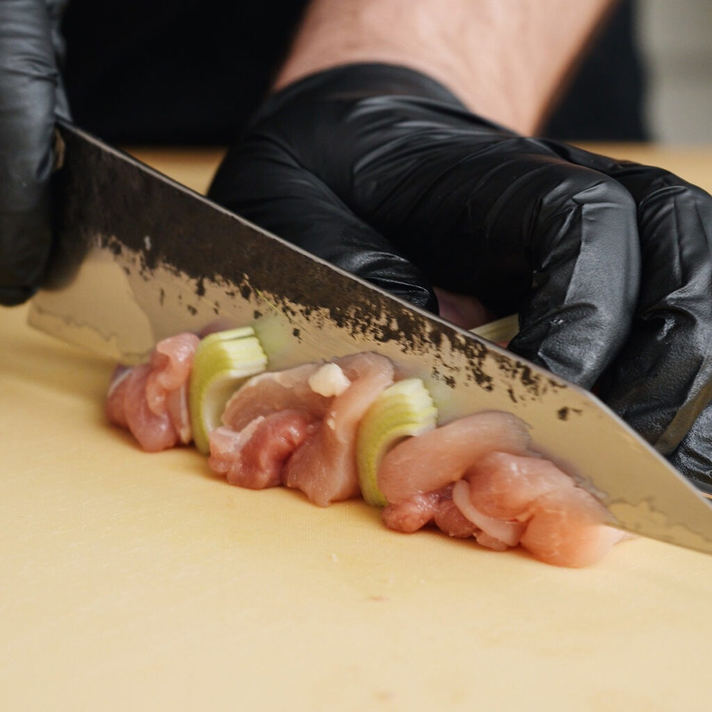 Using a sharp Japanese knife to trim the edges of a raw chicken and leek skewer to ensure an even cook for a homemade yakitori recipe.