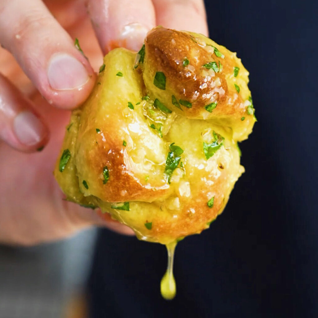 A hand holding a single Dominos copycat garlic knot dripping with warm garlic and herb butter.