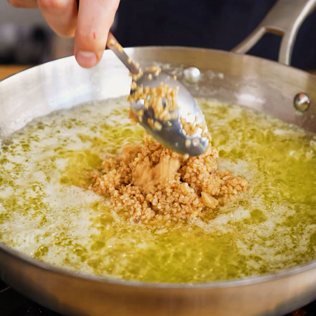 Stirring a spoonful of minced garlic into melted butter and olive oil in a stainless steel skillet.