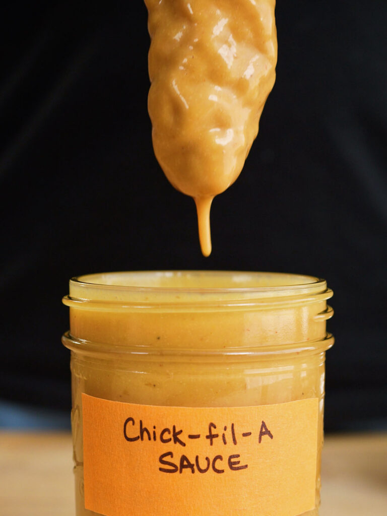 A crispy fried chicken tender being dipped into a jar of thick, sweet mustard and BBQ sauce