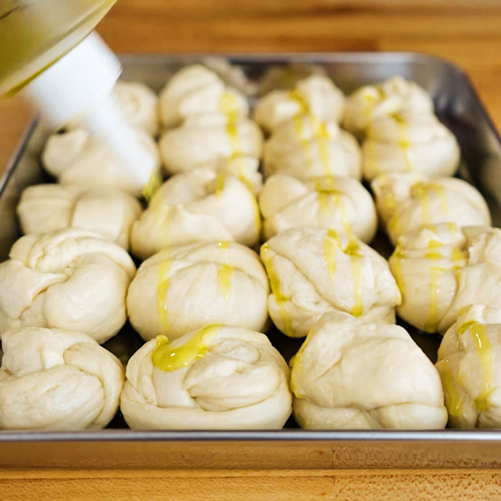 Drizzling olive oil from a squeeze bottle over raw, tied pizza dough on a baking tray to make Dominos copycat garlic knots.