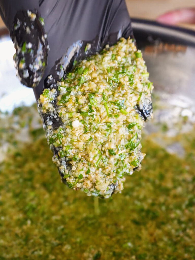 A close-up of a black-gloved hand holding a thick, freshly mixed coating of minced garlic, parsley, and parmesan.