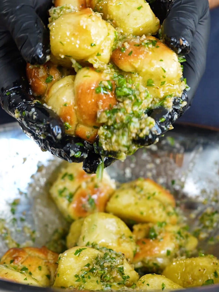Gloved hands tossing hot, baked pizza dough knots in a large metal bowl to thoroughly coat them in a copycat garlic herb sauce.