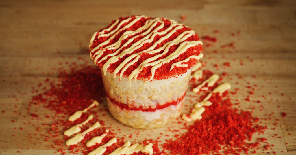 Close-up view of a layered Mexican street corn cup, finished with a spicy chili-lime drizzle and a generous dusting of crushed hot chips.