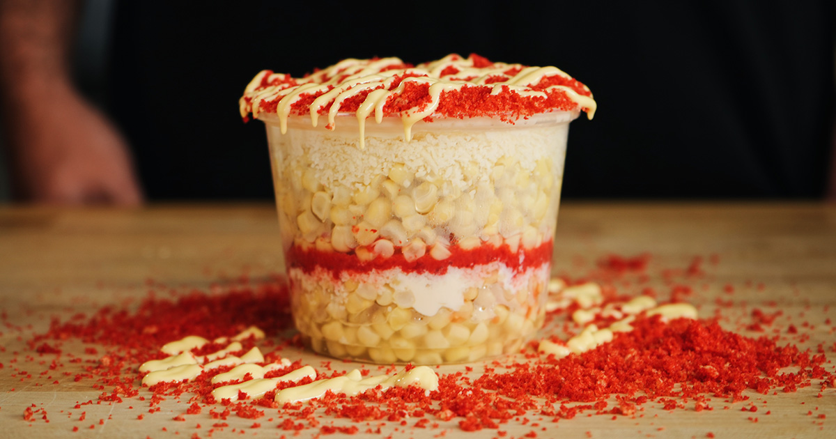 A single serving of authentic Mexican esquites in a plastic cup, featuring visible layers of corn, creamy sauce, cotija cheese, and a crushed Flamin' Hot Cheeto topping. Homemade esquites recipe