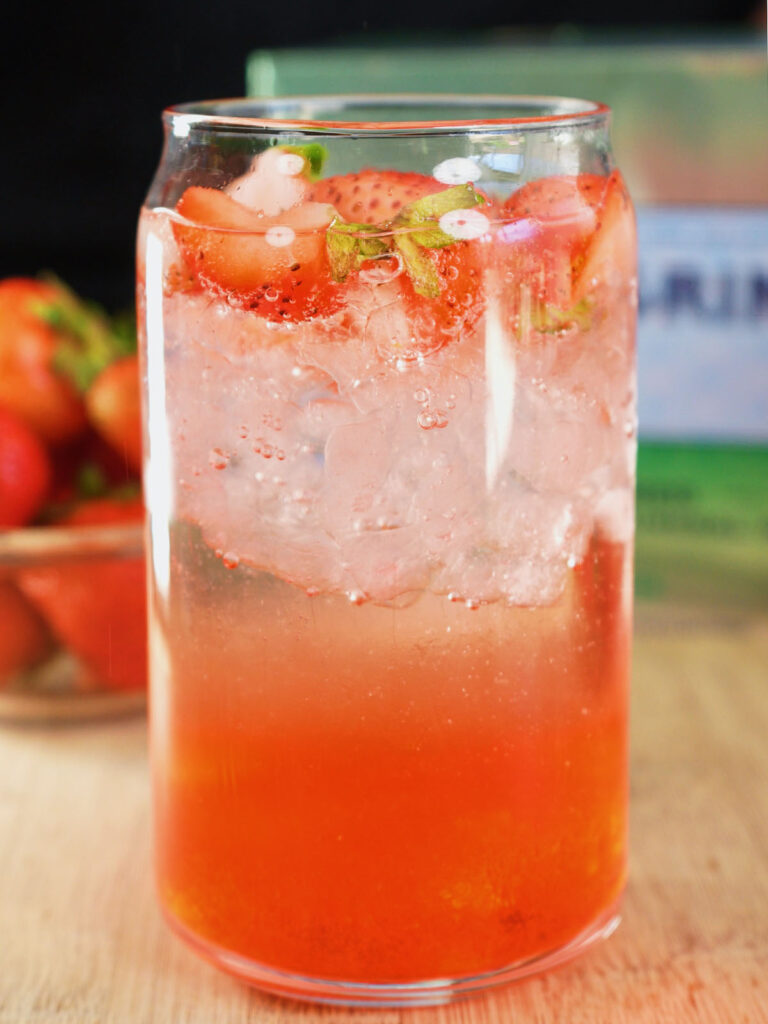 The best homemade strawberry soda recipe in a clear glass, showing a vibrant red fresh fruit syrup at the bottom layered with ice, sparkling water, and diced berries.
