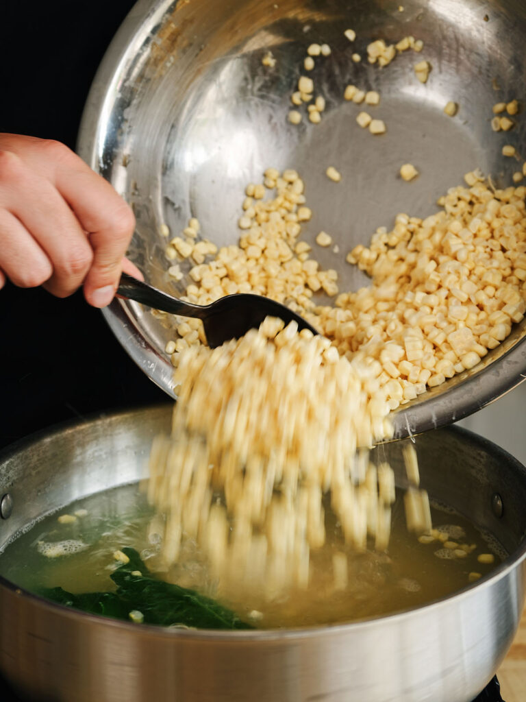 Pouring fresh sweet corn kernels into a pot of boiling water with epazote leaves to prepare the base for an authentic homemade esquites recipe.