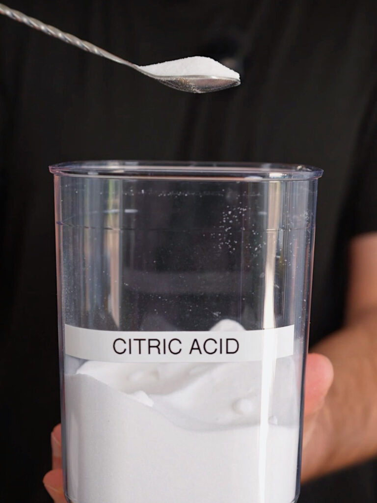 A spoonful of white citric acid powder being added to a clear container to create the sour sugar coating for a Homemade Watermelon Sour Patch recipe.