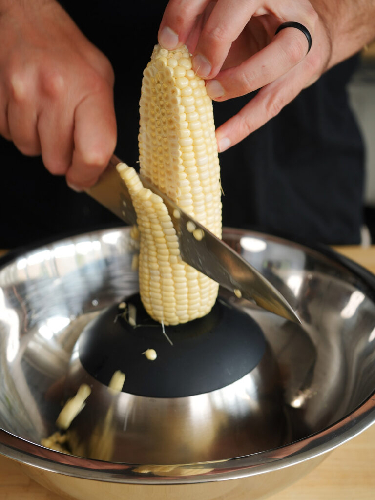 Using a sharp chef's knife to cleanly cut fresh white sweet corn kernels off the cob into a mixing bowl using the inverted bowl preparation technique.