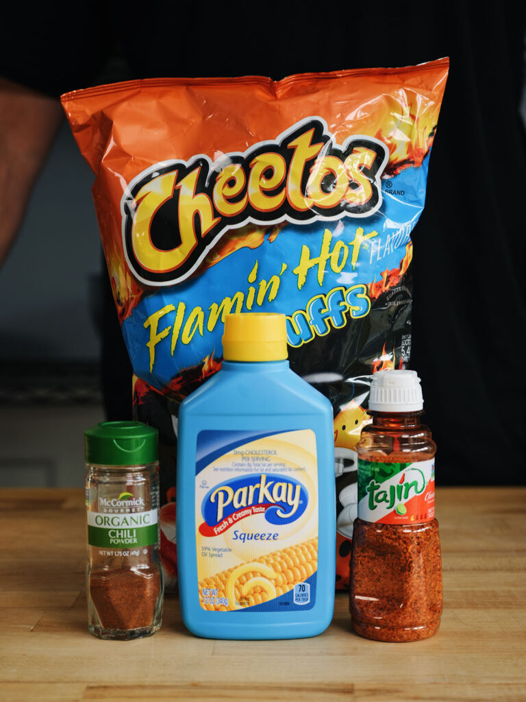 A collection of spicy toppings for Mexican street corn, including a bag of Flamin' Hot Cheetos, chili powder, Tajín seasoning, and squeezable margarine.