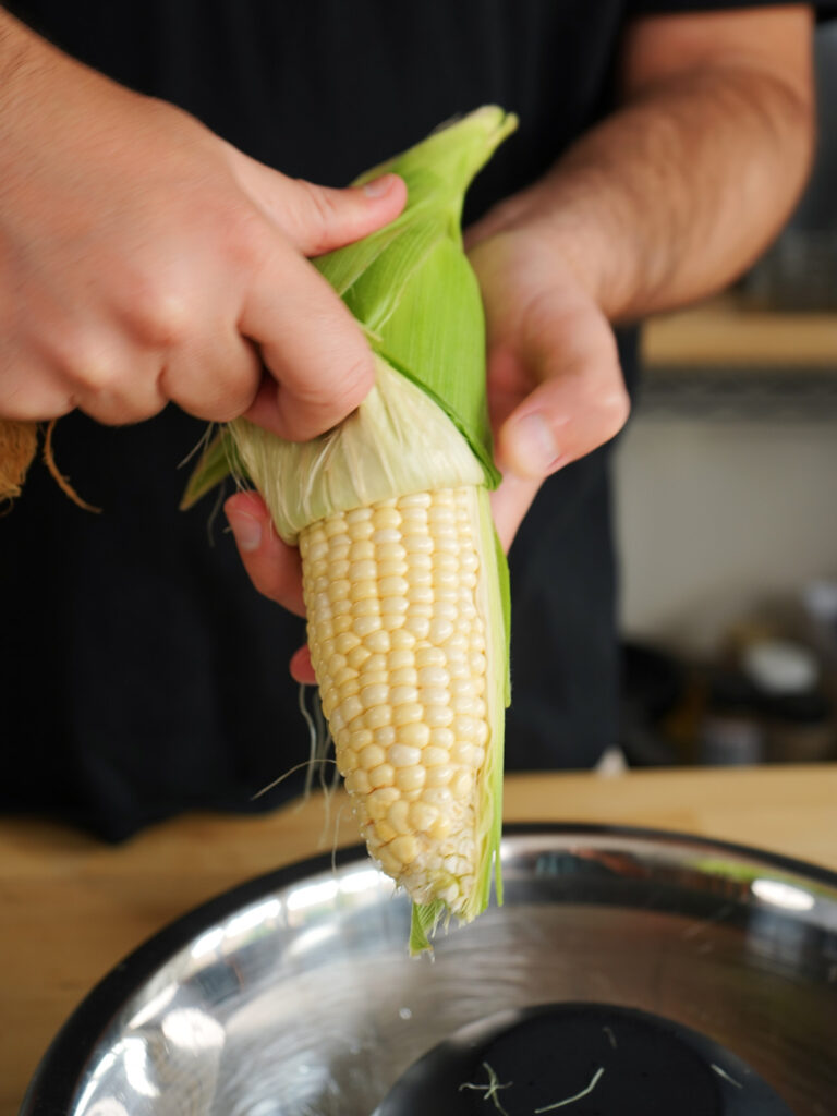Hands peeling the green husk off a fresh ear of sweet corn, demonstrating the preparation step for an authentic street-style corn recipe.