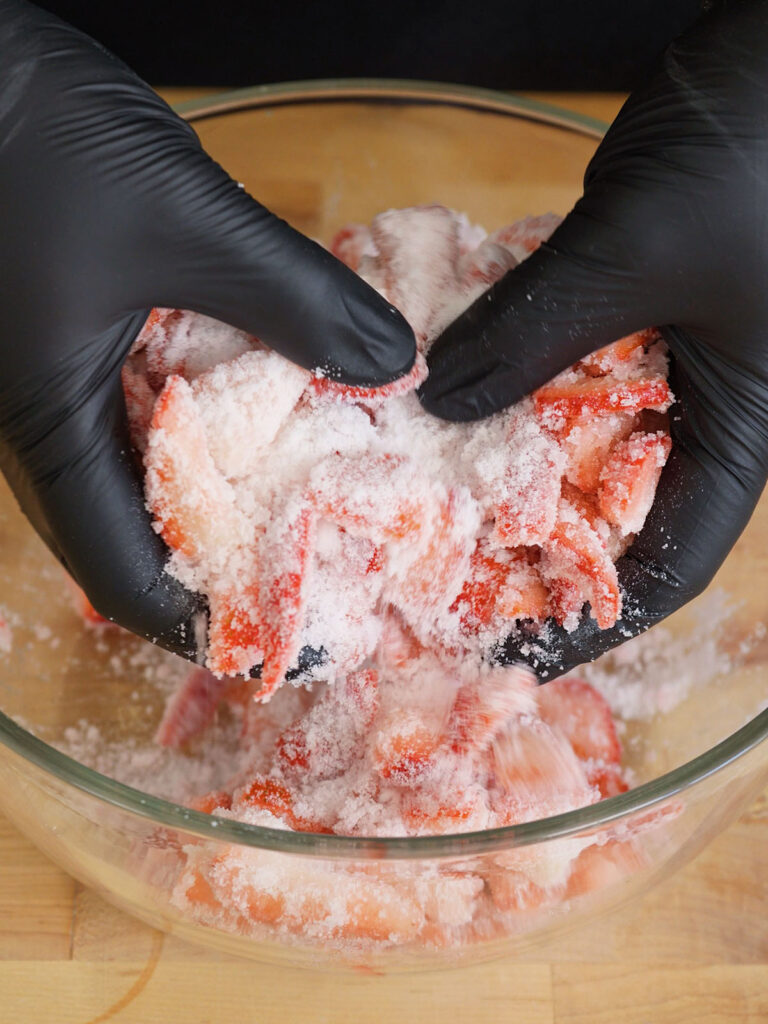 Gloved hands thoroughly mixing fresh diced strawberries with white granulated sugar in a glass bowl to start the maceration process for homemade syrup.