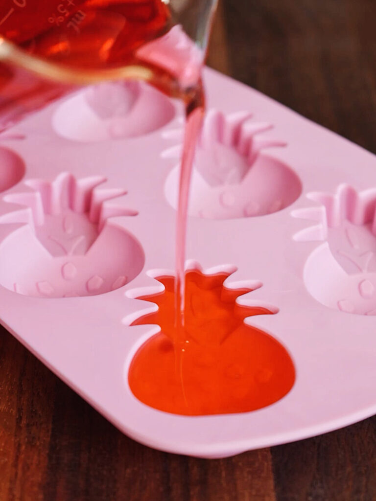 Pouring a hot, red liquid gelatin mixture from a glass beaker into a pink pineapple-shaped silicone mold to set the gummy candies.