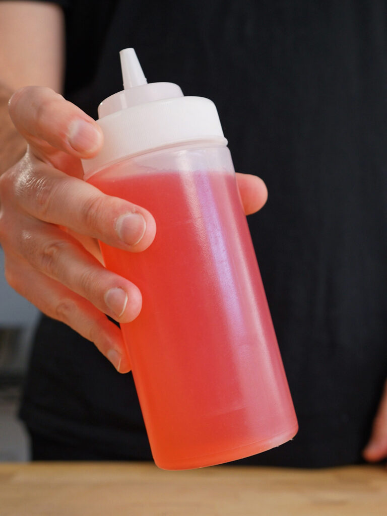 A hand holding a clear squeeze bottle filled with vibrant red, freshly made strawberry syrup ready to be mixed into homemade sodas.