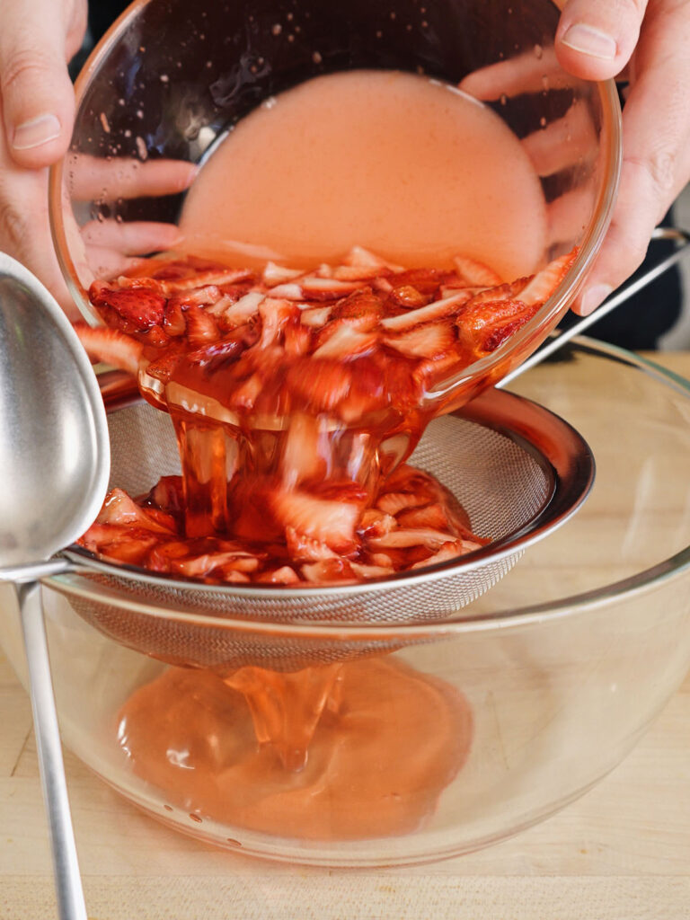 Pouring a mixture of macerated strawberries and syrup through a fine mesh metal strainer into a glass bowl to extract the smooth, fresh fruit syrup.