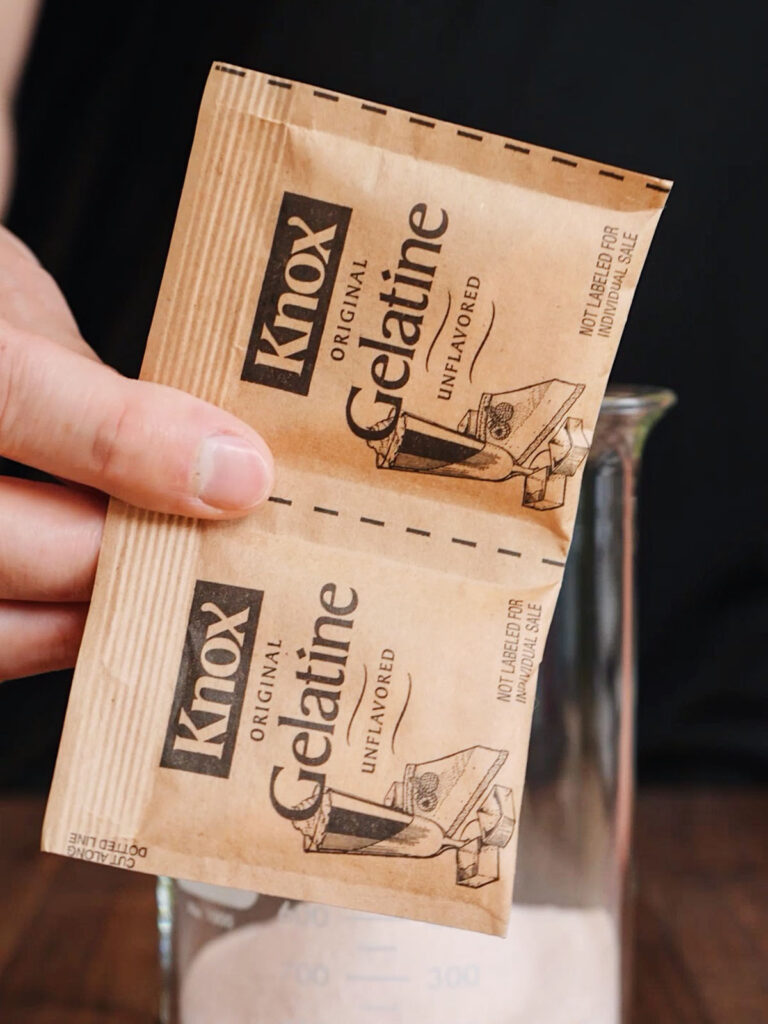 A hand holding two packets of Knox unflavored gelatin over a glass beaker, which creates the chewy, commercial texture for homemade gummy candies.