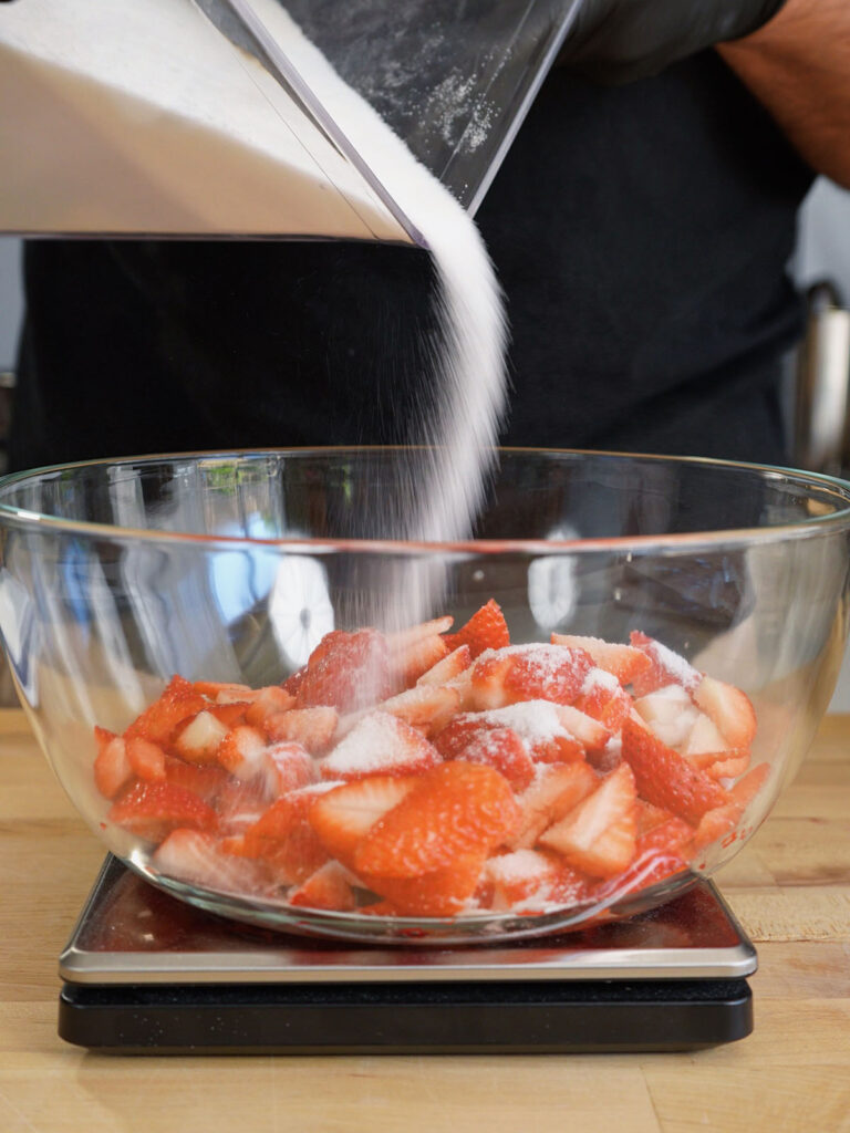 Pouring white granulated sugar over fresh diced strawberries in a clear glass bowl resting on a digital kitchen scale to measure exactly equal parts by mass for a homemade strawberry soda syrup.